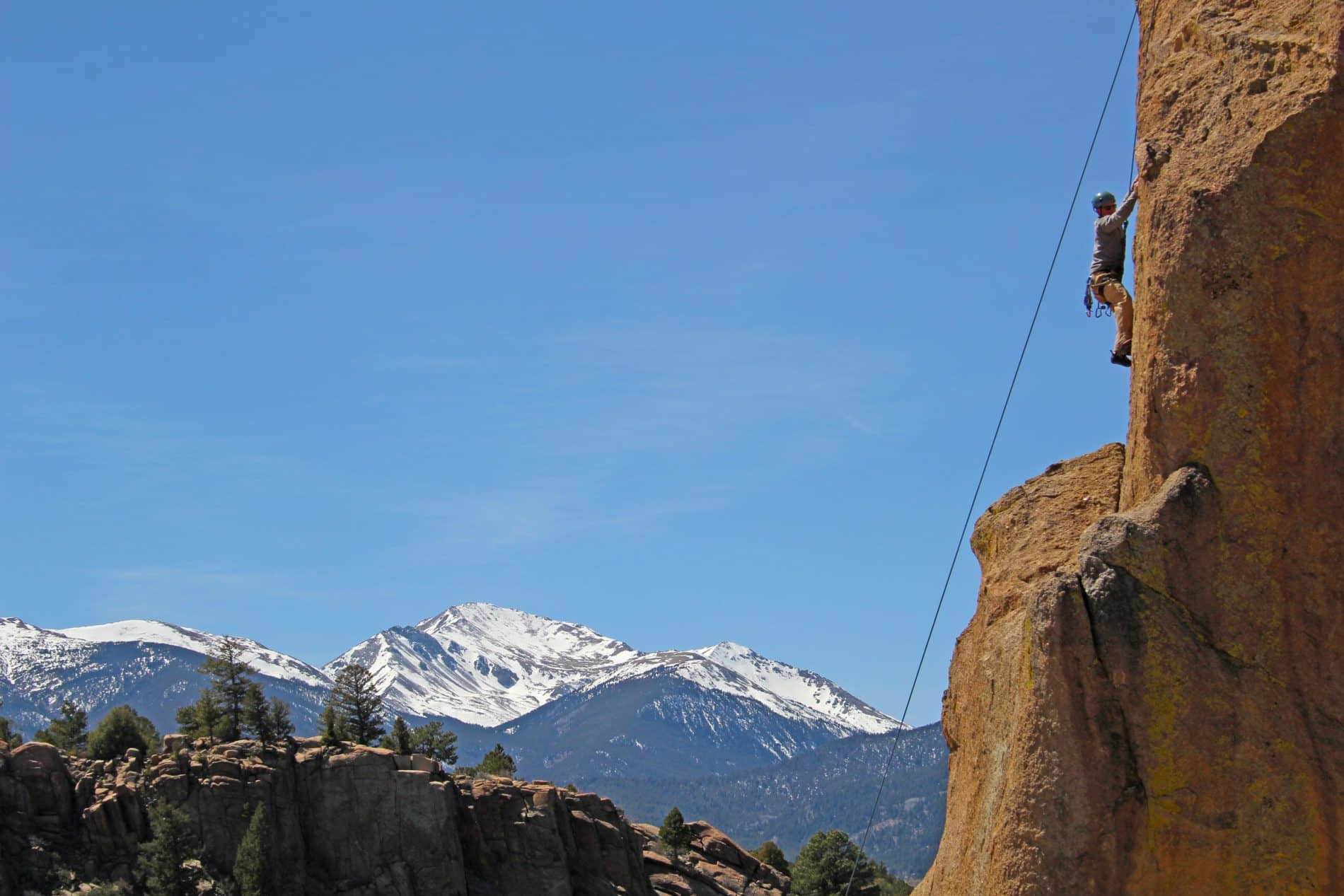 Person climbing a sheer rock face with snow-capped mountains in the background